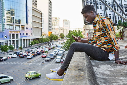 Young man in patterned shirt sitting on roof terrace in the city checking his phone, Maputo, Mozambique
