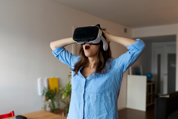 Young woman with mouth open looking through virtual reality simulator while standing in home office