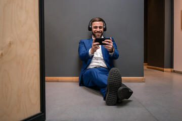 Cheerful male entrepreneur playing video game on smart phone while sitting in corridor at coworking space during break