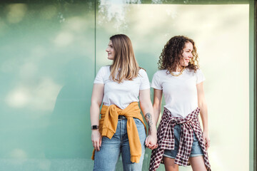 Smiling beautiful women holding hands while looking away against wall