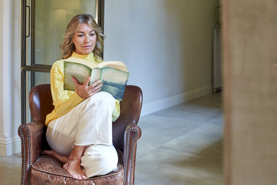 Smiling woman reading book while sitting on armchair at home