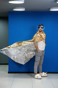Businessman in gold color wrapping paper cape looking away by blue wall at workplace