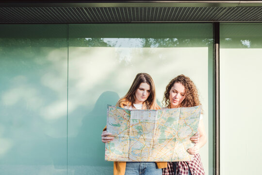 Female friends holding map against wall in city on sunny day