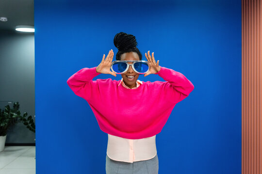 Cheerful creative female professional wearing oversized sunglasses against blue wall at work place