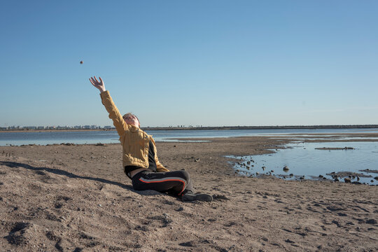 Boy sitting alone on sandy beach and throwing stones