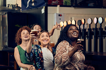 Excited female friends sitting at the counter in a pub watching Tv