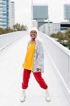 Smiling woman wearing cap with wig standing on bridge in city