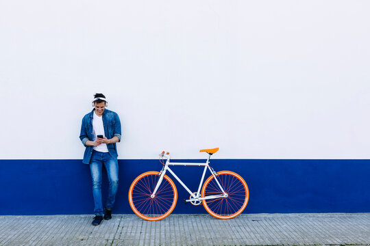 Smiling mature man with mobile phone listening music through wireless headphones by bicycle against wall
