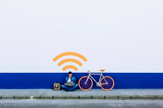 Man in casuals using laptop on sidewalk while sitting at Wi-fi symbol by bicycle against white wall