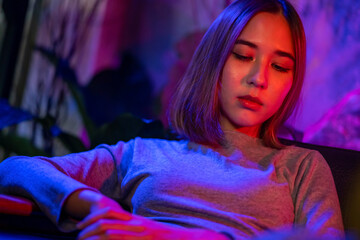 Fototapeta premium Portrait of Loneliness Asian woman sitting on sofa in nightclub with illuminated neon night light with sadness eyes. Beautiful female feeling depressed living alone in multi-colored vibrant lights