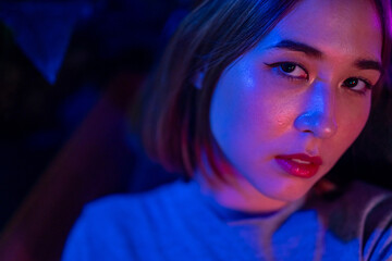 Portrait of Loneliness Asian woman sitting on sofa in nightclub with illuminated neon night light with sadness eyes. Beautiful female feeling depressed living alone in multi-colored vibrant lights