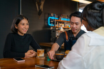 Asian man coffee shop waiter barista serving coffee latte and bakery to woman customer on the table at cafe. Attractive female friend meeting and talking together at restaurant. Small business concept