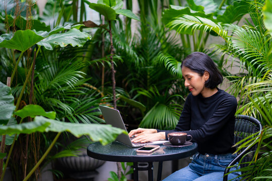 Young beautiful Asian woman freelancer working on laptop computer with drinking a cup of coffee in the garden at home. Attractive female using laptop for remote corporate business or online shopping.