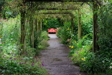 A garden walk through trellises  and greenery that leads to a red bench at the end in the gardens...