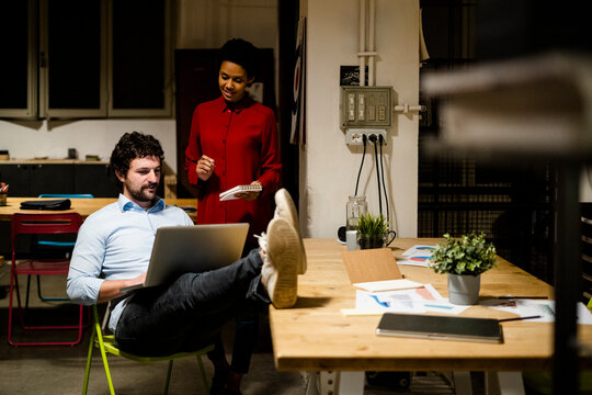 Businesswoman with businessman using laptop in office with feet on desk