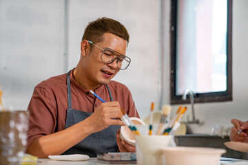Asian LGBTQ guy learning color painting her self-made pottery at home. Confidence male enjoy hobbies and indoors leisure activity handicraft ceramic sculpture and painting workshop at pottery studio