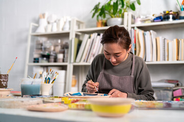 Asian woman learning color painting her self-made pottery at home. Confidence female enjoy hobbies and indoors leisure activity handicraft ceramic sculpture and painting workshop at pottery studio.