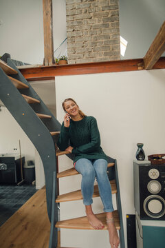 Smiling young woman sitting on stairs at home talking on cell phone