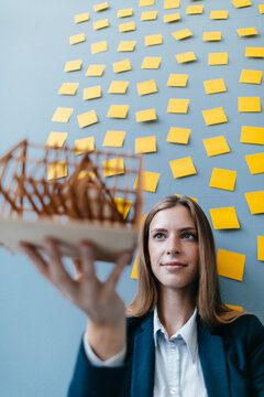 Young businesswoman holding architectural model with yellow sticky notes on the wall behind ger