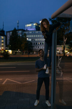 Young man giving girlfriend with cell phone a leg-up in the city at night