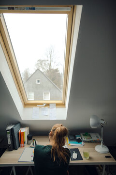 Rear View Of Young Woman Sitting At Desk In Attic