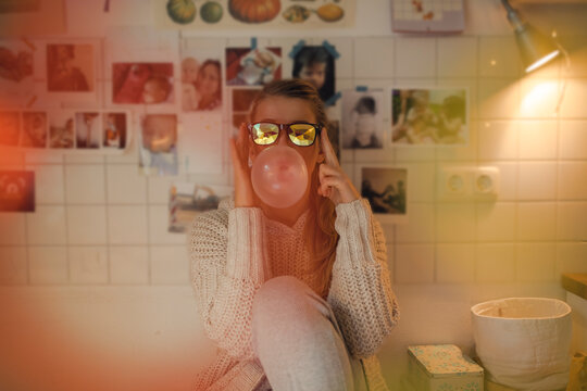 Young woman at home wearing sunglasses making chewing gum bubble