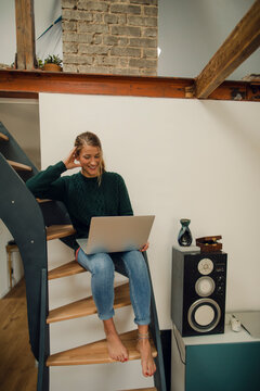 Smiling young woman sitting on stairs at home using laptop
