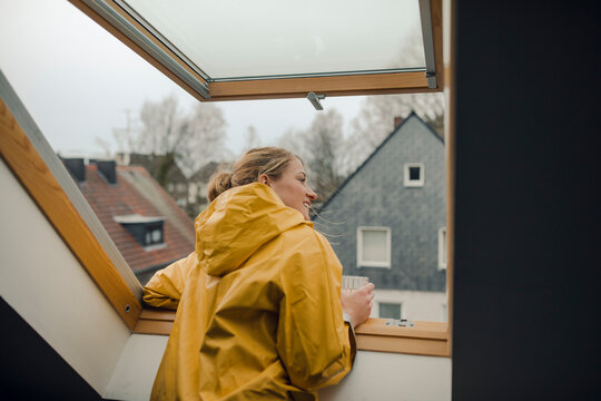 Smiling young woman in raincoat looking out of attic window