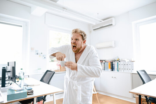 Screaming businessman wearing bathrobe in office putting his hand into coffee cup