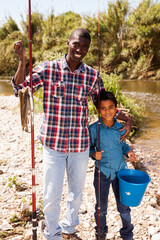 Positive African man and little boy standing near river and holding fishing rod and bucket of catch