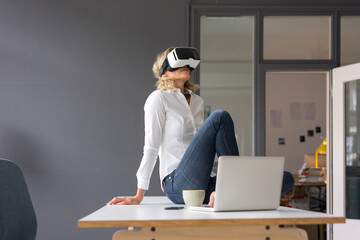 Young businesswoman sitting on desk in office wearing VR glasses