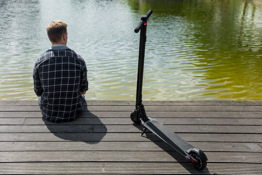 Rear view of yung man with e-scooter sitting on boardwalk