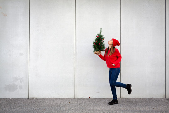 Woman wearing red pullover and wolly hat, holding artificial Christmas tree in front of a wall