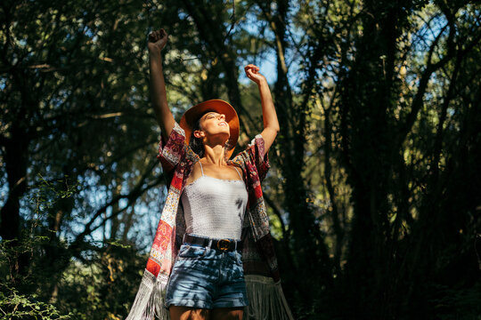 Young woman wearing a brown hat, colorful shirt and white top with closed eyes and arms up feeling the sun in a forest