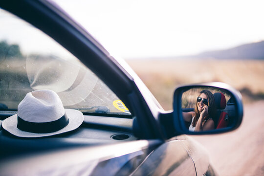 Mirror imag of woman talking on the phone in her car with hat on dashboard