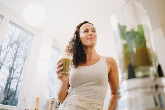 Fit woman standing in kitchen, drinking healthy smoothie