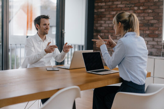 Businessman and woman sitting at desk, working on laptop, pointing with pistol fingers at each other - Powered by Adobe
