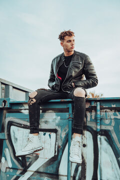 Young man with hands in pockets sitting on metal container full of garffiti against sky