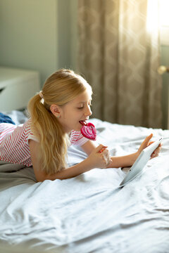 Blond girl lying on bed licking lollipop while using digital tablet