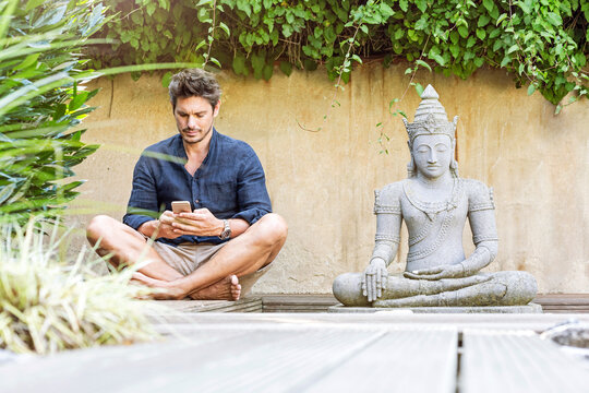 Man sitting cross-legged next to Buddha statue in a Zen garden, using smartphone