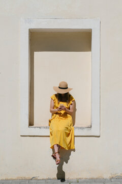 Woman wearing hat using mobile phone while sitting on built structure in city