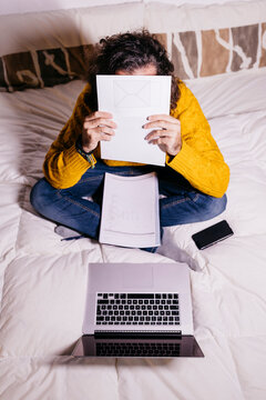 Young man working in bed with laptop covering his face with papers