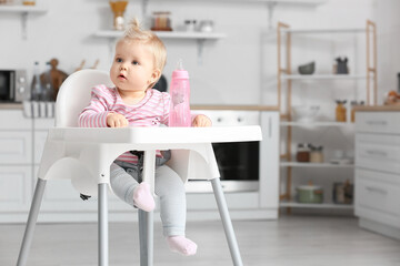 Cute baby girl with bottle of water sitting on high chair in kitchen