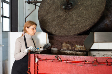 Confident woman engaged in production of olive oil, controlling crushing of olives to fine paste in crusher