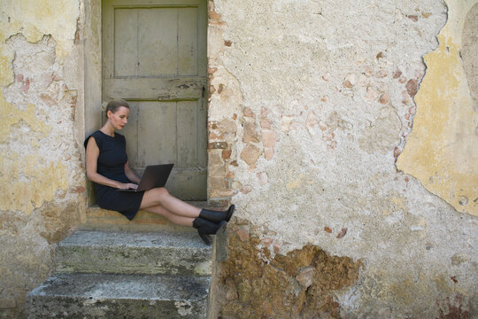 Italy, Tuscany, Monteriggioni, woman sitting at house entrance using laptop