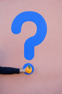Man's hand holding an orange at a wall with question mark
