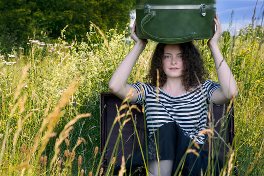 Portrait of young woman sitting in suitcase on a meadow with hatbox on top of her head