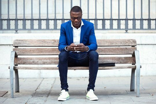 Young businessman wearing blue suit jacket sitting on bench and using smartphone