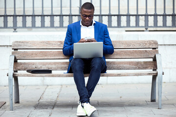 Young businessman wearing blue suit jacket sitting on bench and using laptop