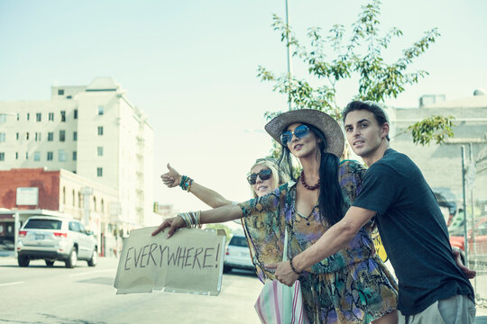 Carefree friends hitchhiking while holding placard with text against sky in city